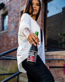 A person wearing a white shirt and black pants is holding a bottle of Sangría Señorial. They are standing on an outdoor staircase with a background of rustic brick walls and windows with reflections. The angle emphasizes the bottle, which is held prominently with a slight tilt towards the viewer.