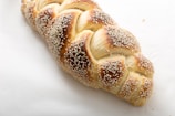 A freshly baked braided bread loaf topped with sesame seeds. The bread has a golden brown crust with a shiny appearance, lying on a light, plain surface.