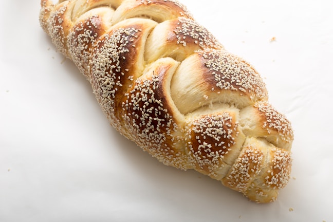 A freshly baked braided bread loaf topped with sesame seeds. The bread has a golden brown crust with a shiny appearance, lying on a light, plain surface.