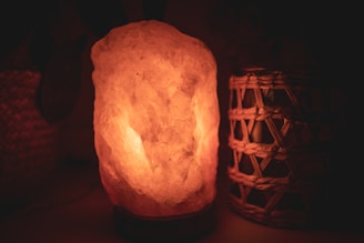 A warm, glowing salt lamp is placed on a surface. The lamp is made from a chunk of salt rock and emits a soothing, amber light. Next to the lamp, there is a wicker or woven container with a candle or small light inside. The background is dimly lit, enhancing the cozy ambiance.