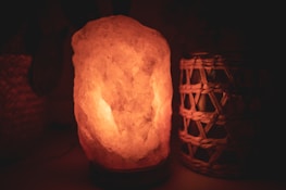 A warm, glowing salt lamp is placed on a surface. The lamp is made from a chunk of salt rock and emits a soothing, amber light. Next to the lamp, there is a wicker or woven container with a candle or small light inside. The background is dimly lit, enhancing the cozy ambiance.