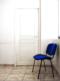 A simple interior space featuring a white door with a white handle set against a tiled floor. Adjacent to the door is a blue chair with black legs, placed near a wall with a window partially visible on the right side.