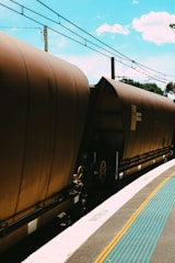 A train with brown freight cars is captured at a railway station. Overhead power lines run parallel to the tracks, and the sky is a bright blue with scattered clouds. A tactile paving strip with blue raised dots runs along the edge of the platform, providing guidance for visually impaired individuals.