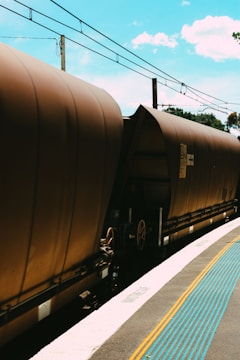 A train with brown freight cars is captured at a railway station. Overhead power lines run parallel to the tracks, and the sky is a bright blue with scattered clouds. A tactile paving strip with blue raised dots runs along the edge of the platform, providing guidance for visually impaired individuals.
