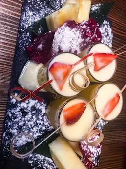 A black rectangular slate serves an artistic arrangement of desserts topped with strawberries and dusted with powdered sugar. The creamy yellow sauce is adorned with vibrant red strawberries arranged on skewers. Surrounding the desserts are maroon gelatin forms and fresh pineapple pieces.