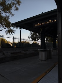 An outdoor transit station with a roofed area featuring columns. There is a digital sign displaying information, and the surrounding area includes trees and bushes behind a metal fence. The scene is captured in low lighting conditions, likely around dusk or dawn.