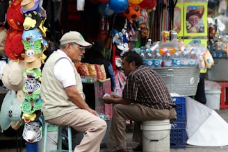 Two men are seated at a bustling market stall. One man is wearing a light vest and cap, sitting on a stool next to a display of colorful hats and masks. The other man, in a checkered shirt, is sitting on a crate beside a cooler filled with bottled beverages. Various items hang and are stacked all around them in a busy outdoor market setting.
