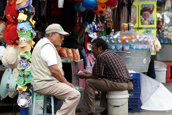 Two men are seated at a bustling market stall. One man is wearing a light vest and cap, sitting on a stool next to a display of colorful hats and masks. The other man, in a checkered shirt, is sitting on a crate beside a cooler filled with bottled beverages. Various items hang and are stacked all around them in a busy outdoor market setting.