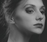 A monochrome portrait of a person with a focused and thoughtful expression. The image captures the side profile of the face, highlighting features such as the eye with well-defined eyeliner, the lips, and the smooth skin texture. The hair is styled back elegantly, with a few strands softly visible. Shadows add depth and contrast, giving the portrait an artistic, classic feel.