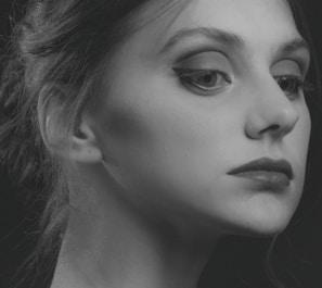 A monochrome portrait of a person with a focused and thoughtful expression. The image captures the side profile of the face, highlighting features such as the eye with well-defined eyeliner, the lips, and the smooth skin texture. The hair is styled back elegantly, with a few strands softly visible. Shadows add depth and contrast, giving the portrait an artistic, classic feel.
