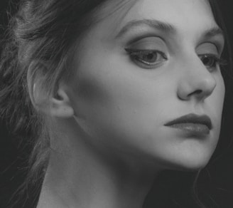 A monochrome portrait of a person with a focused and thoughtful expression. The image captures the side profile of the face, highlighting features such as the eye with well-defined eyeliner, the lips, and the smooth skin texture. The hair is styled back elegantly, with a few strands softly visible. Shadows add depth and contrast, giving the portrait an artistic, classic feel.