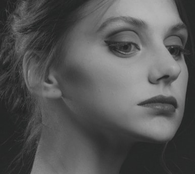 A monochrome portrait of a person with a focused and thoughtful expression. The image captures the side profile of the face, highlighting features such as the eye with well-defined eyeliner, the lips, and the smooth skin texture. The hair is styled back elegantly, with a few strands softly visible. Shadows add depth and contrast, giving the portrait an artistic, classic feel.