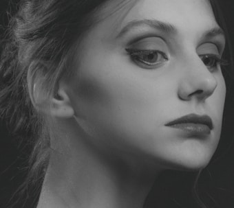A monochrome portrait of a person with a focused and thoughtful expression. The image captures the side profile of the face, highlighting features such as the eye with well-defined eyeliner, the lips, and the smooth skin texture. The hair is styled back elegantly, with a few strands softly visible. Shadows add depth and contrast, giving the portrait an artistic, classic feel.