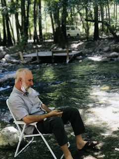 A man sits on a folding chair beside a flowing stream in a tranquil forest setting. He is wearing a face mask under his chin and is looking at his phone. The surrounding area is lush with trees and there is a white vehicle parked in the background.