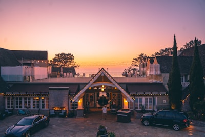 A luxurious house at dusk with a warm, inviting glow from the lights. The foreground features two cars and a small fountain. Beyond the roof, there are trees silhouetted against a colorful evening sky with soft gradient hues.