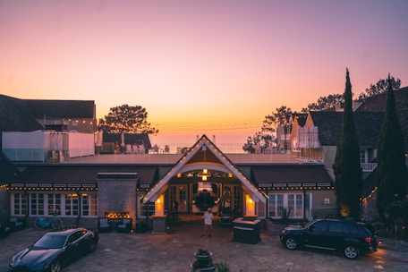 A luxurious house at dusk with a warm, inviting glow from the lights. The foreground features two cars and a small fountain. Beyond the roof, there are trees silhouetted against a colorful evening sky with soft gradient hues.