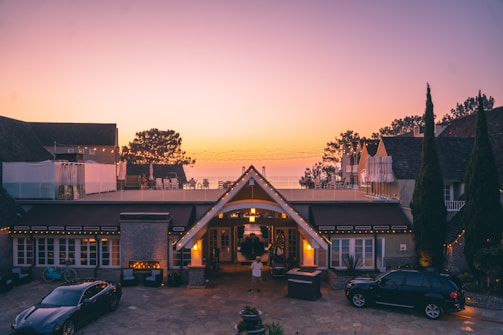 A luxurious house at dusk with a warm, inviting glow from the lights. The foreground features two cars and a small fountain. Beyond the roof, there are trees silhouetted against a colorful evening sky with soft gradient hues.
