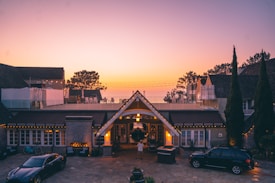 A luxurious house at dusk with a warm, inviting glow from the lights. The foreground features two cars and a small fountain. Beyond the roof, there are trees silhouetted against a colorful evening sky with soft gradient hues.