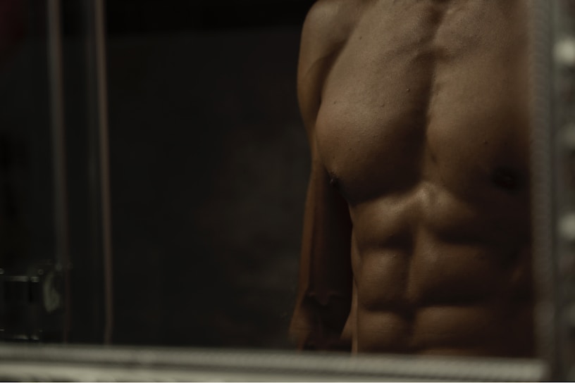A close-up of a male torso with well-defined muscles, including pronounced abs and a prominent chest. The lighting is low, creating strong shadows that accentuate the muscle definition.