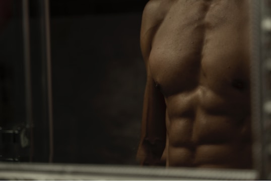 A close-up of a male torso with well-defined muscles, including pronounced abs and a prominent chest. The lighting is low, creating strong shadows that accentuate the muscle definition.