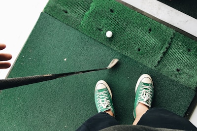 A person wearing green and white sneakers stands on green artificial turf, holding a golf club near a golf ball.