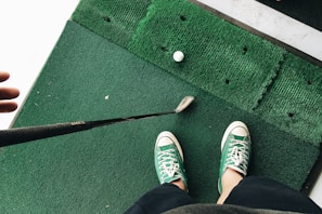 A person wearing green and white sneakers stands on green artificial turf, holding a golf club near a golf ball.