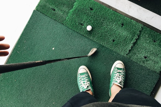 A person wearing green and white sneakers stands on green artificial turf, holding a golf club near a golf ball.
