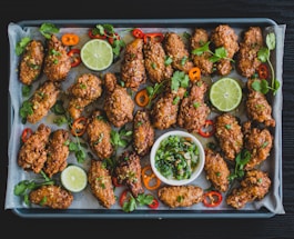 A tray filled with crispy, golden-brown chicken wings garnished with fresh cilantro, sliced red and orange peppers, and lime slices. A small bowl of chopped green onions and herbs sits in the center for dipping.
