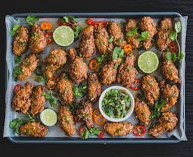 A tray filled with crispy, golden-brown chicken wings garnished with fresh cilantro, sliced red and orange peppers, and lime slices. A small bowl of chopped green onions and herbs sits in the center for dipping.