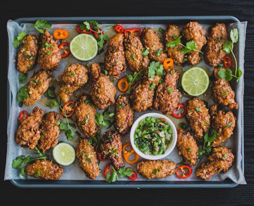 A tray filled with crispy, golden-brown chicken wings garnished with fresh cilantro, sliced red and orange peppers, and lime slices. A small bowl of chopped green onions and herbs sits in the center for dipping.