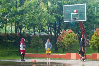 A group of people is gathered on an outdoor basketball court surrounded by lush green trees and bushes. One person is shooting a basketball into the hoop, while two others stand nearby watching and holding a ball. They appear to be engaged in a casual basketball activity.