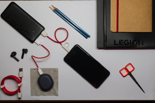 An organized flat lay of various items including a black power bank with a red and white cable attached, a smartphone, a black laptop with a brown notebook tucked inside it, two black stylus pens, a pair of orange-handled scissors, a coiled red USB cable, a small gray pouch, and a couple of blue pencils.