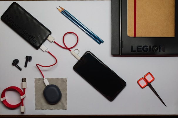 An organized flat lay of various items including a black power bank with a red and white cable attached, a smartphone, a black laptop with a brown notebook tucked inside it, two black stylus pens, a pair of orange-handled scissors, a coiled red USB cable, a small gray pouch, and a couple of blue pencils.
