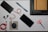 An organized flat lay of various items including a black power bank with a red and white cable attached, a smartphone, a black laptop with a brown notebook tucked inside it, two black stylus pens, a pair of orange-handled scissors, a coiled red USB cable, a small gray pouch, and a couple of blue pencils.