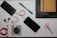 An organized flat lay of various items including a black power bank with a red and white cable attached, a smartphone, a black laptop with a brown notebook tucked inside it, two black stylus pens, a pair of orange-handled scissors, a coiled red USB cable, a small gray pouch, and a couple of blue pencils.
