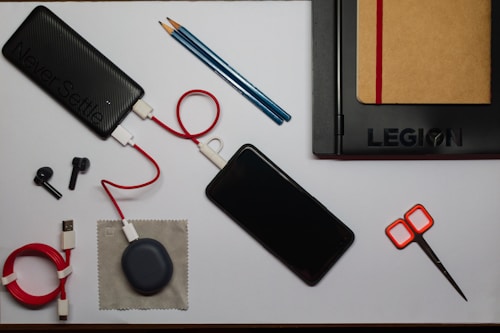 An organized flat lay of various items including a black power bank with a red and white cable attached, a smartphone, a black laptop with a brown notebook tucked inside it, two black stylus pens, a pair of orange-handled scissors, a coiled red USB cable, a small gray pouch, and a couple of blue pencils.