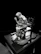 A sculpture of a person seated on a wooden chair with hands clasped in a posture of contemplation or prayer. The figure is draped in a hooded robe, with intricate detailing on the chair and base. The setting appears dramatic, with a strong contrast against the dark background.