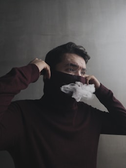 A person stands against a plain background, wearing a dark brown sweater and covering their face with the garment. Wisps of white smoke emanate from their mouth, partially obscured by the fabric.