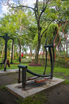 Outdoor exercise equipment surrounded by lush green trees and grass. A person in red clothing is visible in the background. The area has a natural, serene atmosphere with vibrant plant colors.