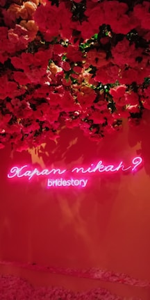 A wall adorned with lush red and pink roses with a neon sign underneath. The sign has pink neon lights displaying the text 'Kapan Nikah? bridestory'. The roses cascade from the top, creating a vibrant floral backdrop.