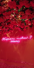 A wall adorned with lush red and pink roses with a neon sign underneath. The sign has pink neon lights displaying the text 'Kapan Nikah? bridestory'. The roses cascade from the top, creating a vibrant floral backdrop.