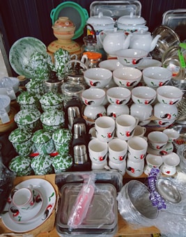 An assortment of kitchenware, including multiple white ceramic bowls and mugs with rooster and floral designs, is displayed. Green enamel cups with white specks and various metal and ceramic teapots, kettles, and dishes are arranged together. The items appear neatly stacked, showcasing a variety of materials and colors.