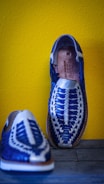 Two handmade, woven leather sandals are displayed against a vibrant yellow wall. The shoes feature a striking blue and white pattern with intricate weaving, showcasing craftsmanship and attention to detail.