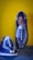 Two handmade, woven leather sandals are displayed against a vibrant yellow wall. The shoes feature a striking blue and white pattern with intricate weaving, showcasing craftsmanship and attention to detail.