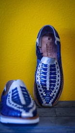 Two handmade, woven leather sandals are displayed against a vibrant yellow wall. The shoes feature a striking blue and white pattern with intricate weaving, showcasing craftsmanship and attention to detail.