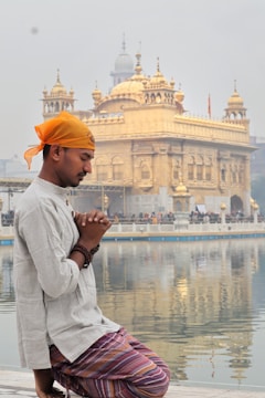 A man wearing a yellow headscarf and traditional attire kneels with hands clasped in prayer by a reflective body of water. Behind him stands a magnificent golden temple, featuring intricate architectural details and multiple domes, under a sky that appears hazy.
