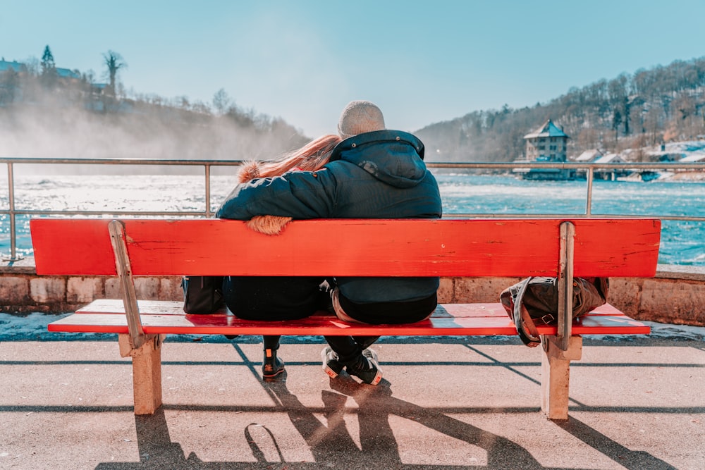 Homme en veste bleue assis sur un banc rouge près d’un plan d’eau pendant la journée