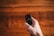 A hand holding a remote control against a wooden surface. The remote has various buttons, some of which include a microphone and power button, and there is a warm, soft focus background.