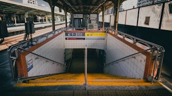 A subway entrance on a train platform has metal railings and tiled walls, leading down a staircase illuminated by natural light. Signs with Japanese writing and directional arrows are displayed above the entrance. The platform is covered with a roof and surrounded by train tracks.