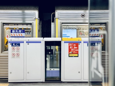 A subway or train station platform with a focus on the safety barriers and doors that separate the platform from the train tracks. The barriers are white with blue and yellow signage, displaying various safety notices and instructions. Behind the barriers, a train with silver and yellow coloring is visible, with some signage and symbols in an East Asian script. The image has a clean and structured appearance with an emphasis on safety and order.